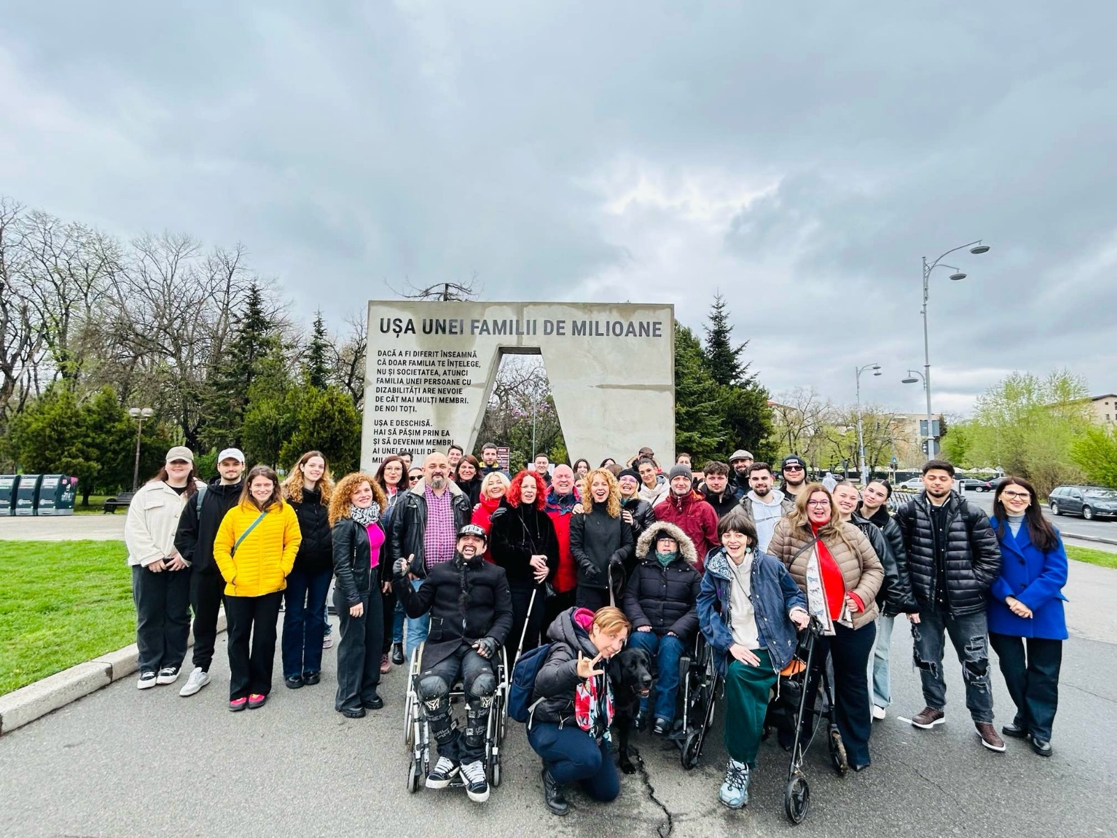 Grup divers de persoane, inclusiv utilizatori de scaun rulant, fotografiat în fața monumentului ‘Ușa unei familii de milioane’ într-un spațiu urban.