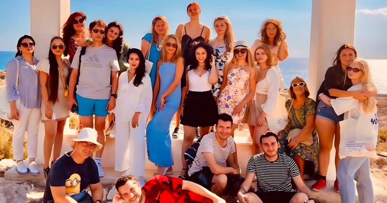 Grup de tineri și tinere zâmbind, fotografiați în aer liber într-un cadru estival, pe fundal cu mare și cer senin, sugerând o activitate de tip Erasmus+, mobilitate sau tabără internațională.