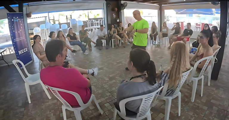 Sesiune de formare informală într-un spațiu deschis, unde un facilitator în tricou verde fosforescent vorbește unui grup de tineri așezați în cerc pe scaune albe din plastic.