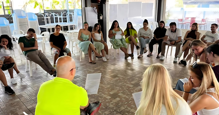 Grup de tineri așezați în cerc pe scaune, participând la o sesiune de discuții sau activitate de formare într-un spațiu interior cu lumină naturală, fiecare ținând foi în mâini.