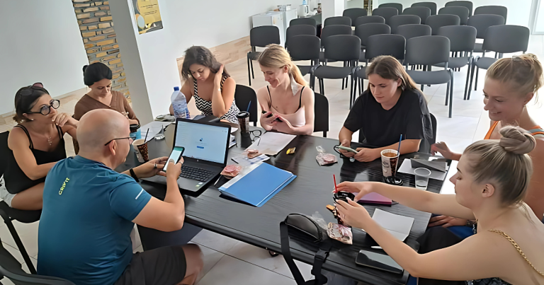 Un grup de participanți așezați la o masă într-o sală de conferințe, lucrând pe telefoane și laptopuri, concentrați în cadrul unui atelier practic.