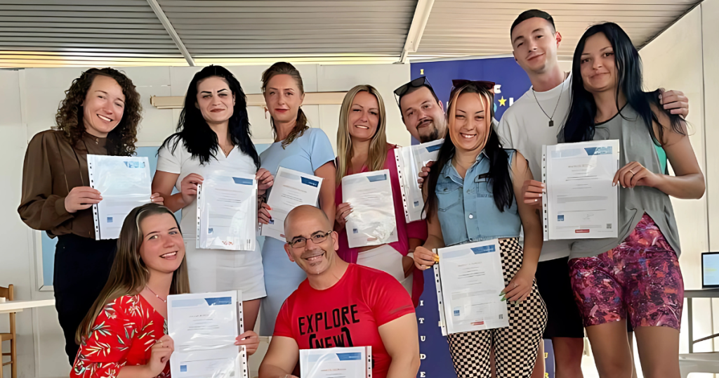 Un grup de participanți zâmbitori țin în mâini certificate de participare, fotografiați într-un spațiu de formare informal, în fața unui banner european.