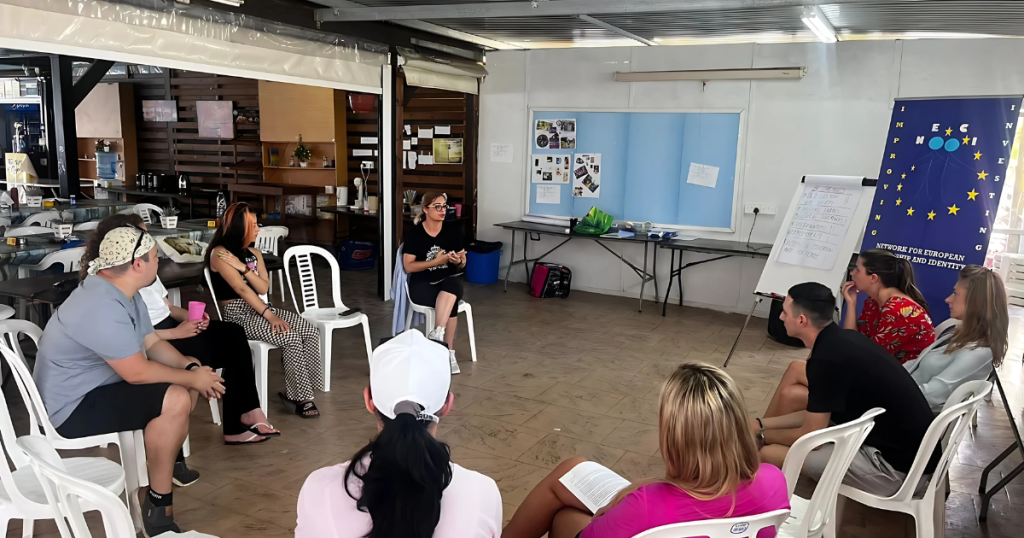 Atelier de formare în desfășurare, cu participanți așezați în cerc ascultând un vorbitor, într-un spațiu informal decorat cu materiale vizuale și un flipchart.