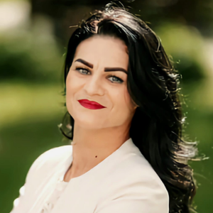 Femeie cu păr brunet ondulat, ruj roșu și sacou alb, privind în cameră, pe fundal natural verde.