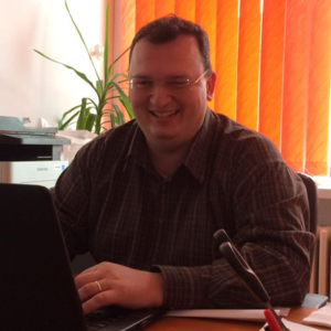 Bărbat zâmbitor, purtând o cămașă în carouri, lucrând la un laptop, într-un birou cu perdele portocalii.