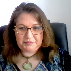 Femeie cu ochelari și păr șaten, purtând un colier cu pandantiv verde, fotografiată pe un scaun de birou.