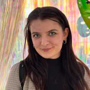 Femeie tânără cu păr brunet și lung, purtând cercei rotunzi și ținută în carouri, fotografiată pe un fundal colorat și decorativ.