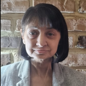 Femeie cu păr negru și drept, tuns scurt, purtând un sacou gri, fotografiată în fața unui perete de cărămidă.