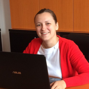 Femeie zâmbitoare stând la birou, purtând un cardigan roșu, lucrând la un laptop ASUS