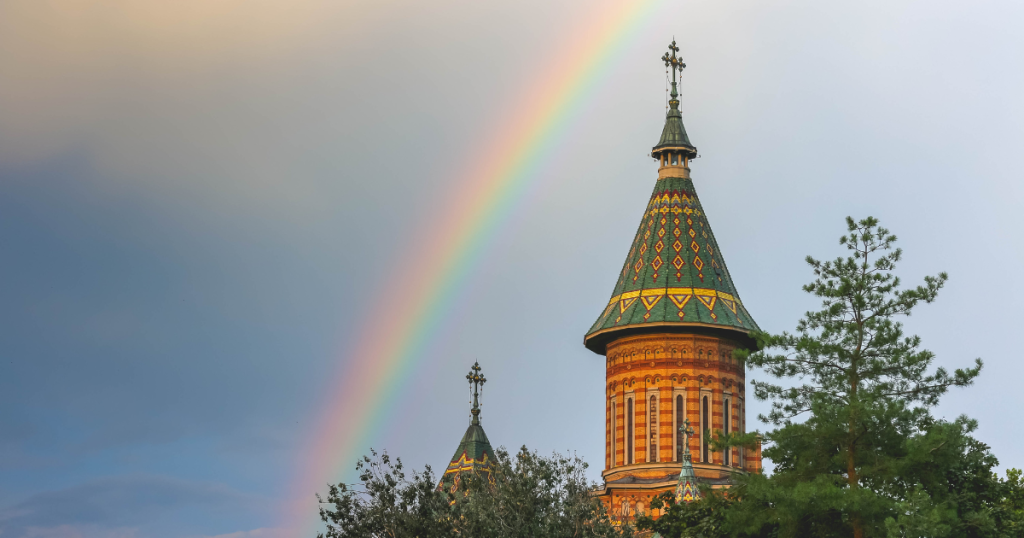 Turnul unei biserici ortodoxe cu acoperiș verde ornamentat, surprins pe fundalul unui cer parțial înnorat, traversat de un curcubeu viu colorat. În prim-plan se văd copaci verzi care încadrează clădirea.