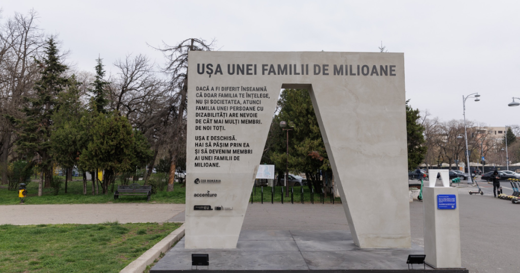 Monument urban în formă de ușă, intitulat ‘Ușa unei familii de milioane’, amplasat într-un spațiu public verde. Textul inscripționat promovează incluziunea persoanelor cu dizabilități în familie și societate. Lângă monument se află o placă informativă și se văd trotinete parcate, bănci și trecători în fundal.
