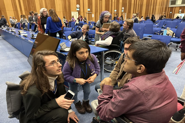 Grup de persoane cu dizabilități în dialog activ într-o sală de conferință internațională, unii în scaune rulante, alții folosind bastoane albe. Atmosferă de incluziune și schimb de idei.