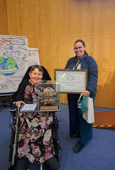 O femeie în scaun rulant ține un trofeu și o diplomă Discovery Awards, alături de o altă femeie care zâmbește și ține un certificat. În fundal, un panou cu desene și text legat de incluziune și schimbări climatice.