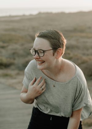 Persoană tânără cu ochelari, râzând în aer liber, pe un fundal natural.